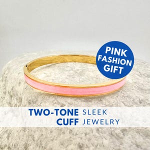 May include: A gold-tone cuff bracelet with a pink enamel center band. The bracelet has the text "PINK FASHION GIFT" on a blue circle and "TWO-TONE SLEEK CUFF JEWELRY" in blue text.