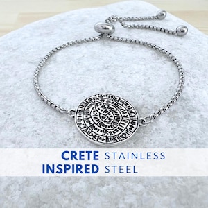 Op de afbeelding: Een zilverkleurige roestvrijstalen armband met een ronde, gegraveerde hanger. De hanger heeft een ingewikkeld ontwerp. De armband heeft een schuifsluiting voor verstelbare maatvoering. De tekst "CRETE INSPIRED STEEL" staat eronder.