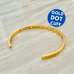 May include: A gold-colored cuff bracelet with small, evenly spaced dot details. The open-ended design features a smooth, polished finish. The text "GOLD DOT CUFF" is in a blue circle in the background.