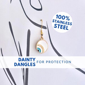 May include: Gold dangle earrings with a white and blue eye design. The earrings are made of 100% stainless steel. "DAINTY DANGLES FOR PROTECTION" is written on the white background.