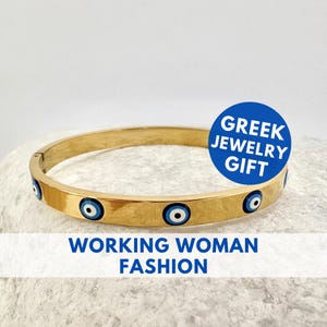 May include: Gold-tone bangle bracelet featuring blue and white evil eye charms. The bracelet has a hinged clasp and is inscribed with the text "GREEK JEWELRY GIFT" and "WORKING WOMAN FASHION".