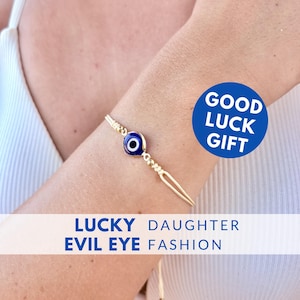 Può includere: Bracciale dorato con un ciondolo a forma di occhio blu e bianco. Il bracciale è indossato su un polso e presenta il testo "GOOD LUCK GIFT" e "LUCKY DAUGHTER EVIL EYE FASHION".
