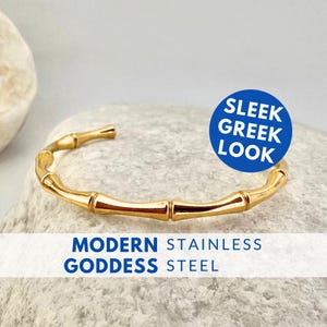 Op de afbeelding: Een goudkleurige roestvrijstalen armband met een bamboe-ontwerp. De armband is open en rust op een grijze steen. Tekst op de afbeelding luidt "Sleek Greek Look" en "Modern Stainless Goddess Steel."