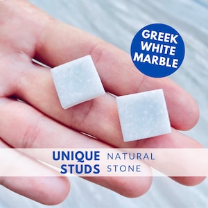 Könnte beinhalten: Zwei quadratische Ohrstecker aus hellem Marmor. Die Ohrringe werden in einer Hand gehalten und sind mit "UNIQUE STUDS NATURAL STONE" und "GREEK WHITE MARBLE" in einem blauen Kreis beschriftet.