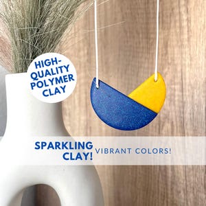 Puede incluir: Un collar hecho con arcilla polimérica de alta calidad. El colgante tiene forma de semicírculo con un diseño azul y amarillo. La parte azul es brillante y la parte amarilla es sólida. El texto "HIGH-QUALITY POLYMER CLAY" está en un círculo blanco.