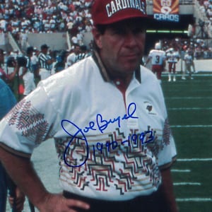 May include: A man wearing a white shirt with a geometric pattern and a red baseball cap with "PHOENIX CARDINALS" written on it. He is standing on a green field with a crowd of people in the background. His signature, "Joe Bugel 1990-1992", is written in blue ink on the shirt.