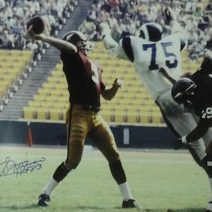 May include: A black and white photo of a football player throwing a football. The player is wearing a brown and gold uniform with the number 4 on the front. The player is surrounded by a crowd of people in the background. The photo is signed by the player with the name "Boomer Esiason" and the date "11/8/83".