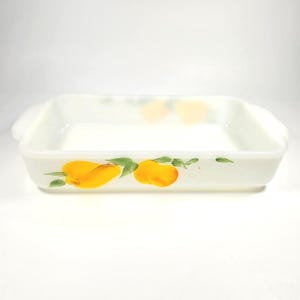 Puede incluir: Una fuente de horno rectangular de vidrio lechoso blanco con asas laterales integradas. El exterior presenta un diseño pintado de melocotones amarillos y hojas verdes en la parte frontal, con un acabado liso y brillante y un interior profundo.