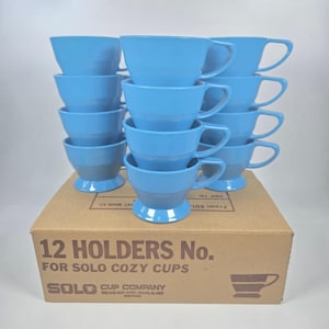 Könnte beinhalten: Ein Set von zwölf hellblauen Solo Cozy Cups, gestapelt auf einem Karton. Die Tassen haben einen Sockel und einen Henkel. Auf der Box steht "12 HOLDERS No. FOR SOLO COZY CUPS" und das Logo der Solo Cup Company.