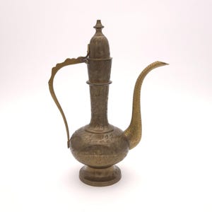May include: A vintage brass pitcher with an ornate design. The pitcher features a long, curved spout, a decorative handle, and a detailed floral pattern etched across its surface. The piece has a bulbous base and a tall, slender neck with a lid.