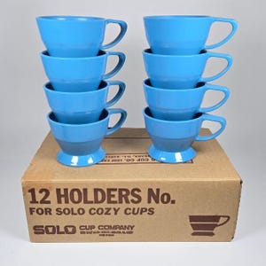 Könnte beinhalten: Ein Satz von zwölf hellblauen Kunststoff-Becherhaltern, die auf einem Karton gestapelt sind. Die Becher haben ein klassisches Design mit Henkel und Sockel. Auf der Box steht "12 HOLDERS No. FOR SOLO COZY CUPS".