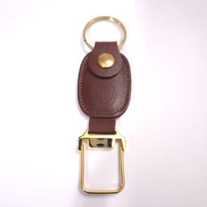 May include: A brown leather keychain with a gold-colored key ring and a gold-colored rectangular clasp. The leather portion features a circular emblem and is secured with a gold-colored rivet.