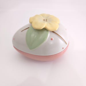 May include: A decorative ceramic egg-shaped container. The container is primarily white with a pink base. It features a yellow flower and a green leaf on top, with pink polka dots and brown lines for added detail.