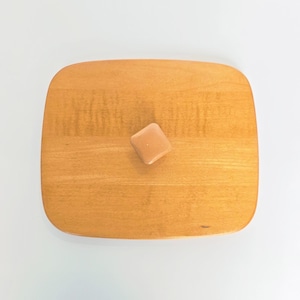 May include: A light brown, square-shaped object rests on a wooden cutting board. The board is rectangular with rounded corners and a light wood grain. The object appears to be a small, square-shaped food item.
