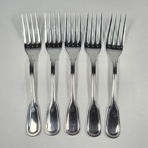 May include: Five silver forks with tines and elongated handles are arranged in a row. The forks have a classic design, with a polished finish that reflects light. The tines are evenly spaced, and the handles have a subtle curve.