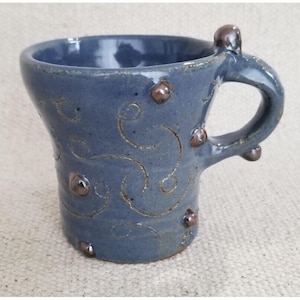 May include: A handmade ceramic mug in a dusty blue hue. The mug features a unique handle design and is adorned with brown, raised embellishments and etched swirl patterns. This stoneware mug is perfect for coffee or tea.