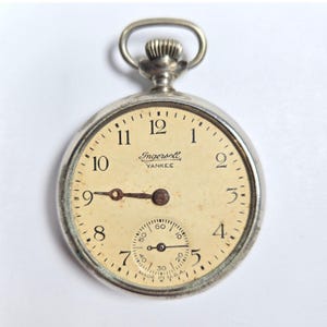 May include: A silver-tone Ingersoll Yankee pocket watch with a cream-colored dial, black Arabic numerals, and a sub-dial for seconds. The watch features a winding crown at the top and the text Ingersoll YANKEE and MADE IN U.S.A. on the face.