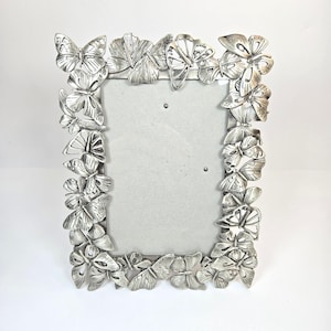 May include: A rectangular picture frame with a metallic silver finish, featuring a decorative border composed of numerous sculpted butterflies of varying sizes and wing positions that completely surround the central rectangular display area.