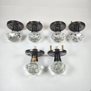 May include: A set of six cabinet knobs featuring clear, multi-faceted glass heads shaped like diamonds. Each knob has a dark bronze-toned circular metal base with mounting hardware, including small screws and pins visible on the back plates.