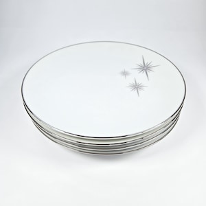 May include: A stack of white ceramic plates with a silver rim and a starburst design in gray. The plates are round and appear to be new. The starburst design is located on the upper right side of the plates.