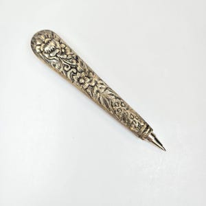 May include: A metallic pen with a tapered, rounded top and a pointed tip. The barrel features an intricate, embossed floral pattern with a tarnished silver finish, showcasing detailed petals, leaves, and swirling vines across the entire surface.