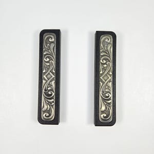 May include: Two black rectangular decorative panels with silver-colored, ornate floral scrollwork. The panels have a rectangular shape with rounded corners. The silver design is centered on the black background.