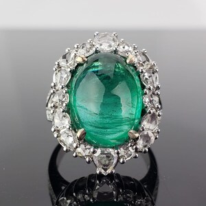 May include: A silver ring with a large, oval-shaped emerald gemstone surrounded by a halo of smaller, round diamonds.