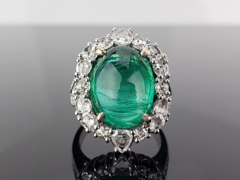 May include: A silver ring with a large, oval-shaped emerald gemstone surrounded by a halo of smaller, round diamonds.