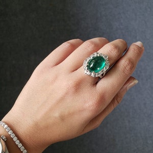May include: A ring with a large, oval-shaped emerald gemstone surrounded by smaller diamonds. The ring is set in white gold.