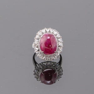 Puede incluir: Un anillo de plata con una gran piedra de rubí ovalada rodeada de piedras más pequeñas y transparentes.