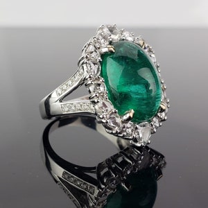 May include: A silver ring with a large, oval-shaped emerald gemstone surrounded by smaller diamonds. The ring has a split shank with diamonds set along the band.