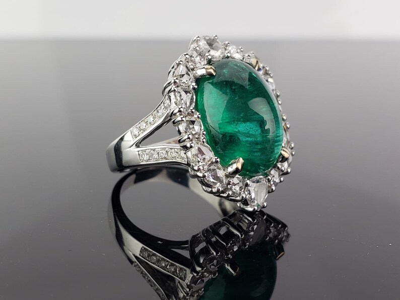 May include: A silver ring with a large, oval-shaped emerald gemstone surrounded by smaller diamonds. The ring has a split shank with diamonds set along the band.