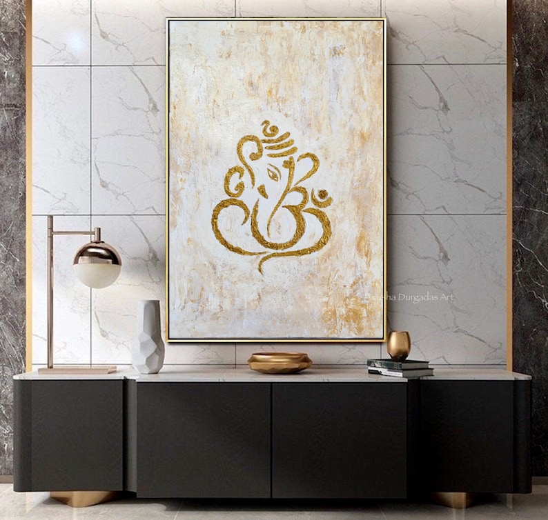 Op de afbeelding: Een abstract schilderij in goud en wit van de hindoe-god Ganesha, met het Om-symbool ge&iuml;ntegreerd in het ontwerp. Het schilderij is ingelijst en hangt aan een muur in een modern interieur.