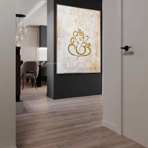 Op de afbeelding: Een gouden Ganesha-symbool op een wit en gouden, gestructureerd canvas schilderij. Het schilderij is ingelijst en hangt aan een muur in een modern huis.