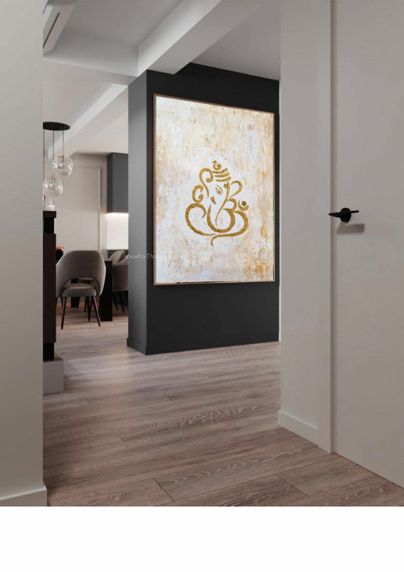 Op de afbeelding: Een gouden Ganesha-symbool op een wit en gouden, gestructureerd canvas schilderij. Het schilderij is ingelijst en hangt aan een muur in een modern huis.