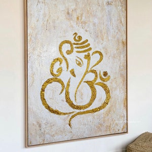 Op de afbeelding: Een wit canvas schilderij met een gouden Ganesha-ontwerp. Het schilderij is ingelijst in een licht houten lijst.