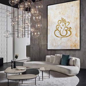 Op de afbeelding: Een abstract wit en gouden schilderij van de hindoe-god Ganesha, met een gouden lijst, hangt aan een grijze muur in een moderne woonkamer. De kamer heeft een witte bank, een rond tapijt en een set van drie salontafels.