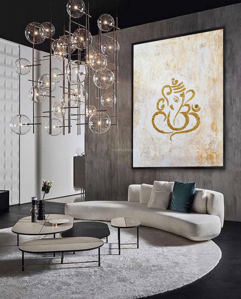 Op de afbeelding: Een abstract wit en gouden schilderij van de hindoe-god Ganesha, met een gouden lijst, hangt aan een grijze muur in een moderne woonkamer. De kamer heeft een witte bank, een rond tapijt en een set van drie salontafels.