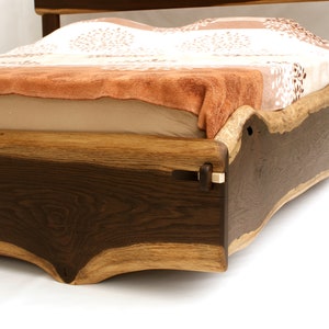 May include: A wooden bed frame with a natural wood finish. The frame is made from a single piece of wood with a live edge. The bed is covered with a white and brown patterned blanket.