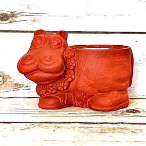 May include: A terracotta-colored hippo planter. The hippo has a detailed face and a floral garland around its neck. The planter has a large opening for plants. The hippo is sitting on a white wooden surface.