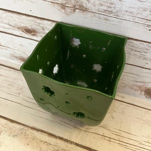 Peut inclure: Un récipient en céramique vert foncé de forme carrée avec des motifs floraux découpés. Le récipient a une finition brillante et est posé sur un fond en bois clair.