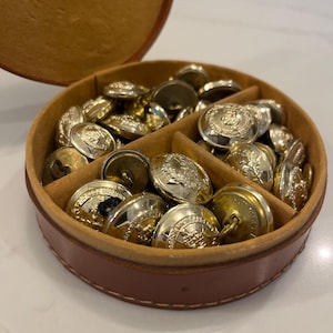 May include: A selection of shiny, gold-coloured buttons with detailed embossed designs, housed in a round, brown leather case with a divided interior. The buttons are of various sizes and shapes.