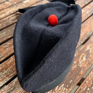 Könnte beinhalten: Ein schwarzer Wollhut mit einem roten Bommel oben. Der Hut hat ein schwarzes Band, das um ihn gebunden ist.