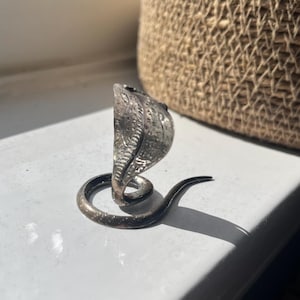 Puede incluir: Un anillo de metal plateado con forma de serpiente cobra con la capucha desplegada. La banda del anillo forma el cuerpo enroscado de la serpiente, mientras que la cabeza y la capucha están posicionadas erguidas, con escamas y marcas grabadas.