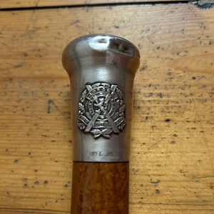 May include: Close-up of a vintage walking cane. The handle is silver-toned with an emblem and text. The shaft is a warm brown colour. The cane is resting on a wooden surface.