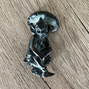 May include: A black metal door knocker shaped like a goblin sitting on a mushroom. The goblin has a mischievous expression and is holding a flower in its hand.