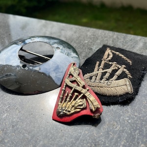 Puede incluir: Tres broches decorativos con gaitas. Una broche es plateada, otra es roja y dorada, y la última es negra con bordados dorados.