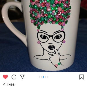 May include: White ceramic mug with a black line drawing of a woman with glasses and a pink and green rhinestone afro. The mug is decorated with pink and green rhinestones.