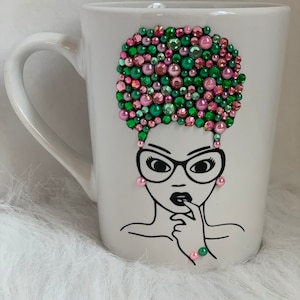 May include: White ceramic mug with a black line drawing of a woman with glasses and an afro. The afro is decorated with pink and green rhinestones.