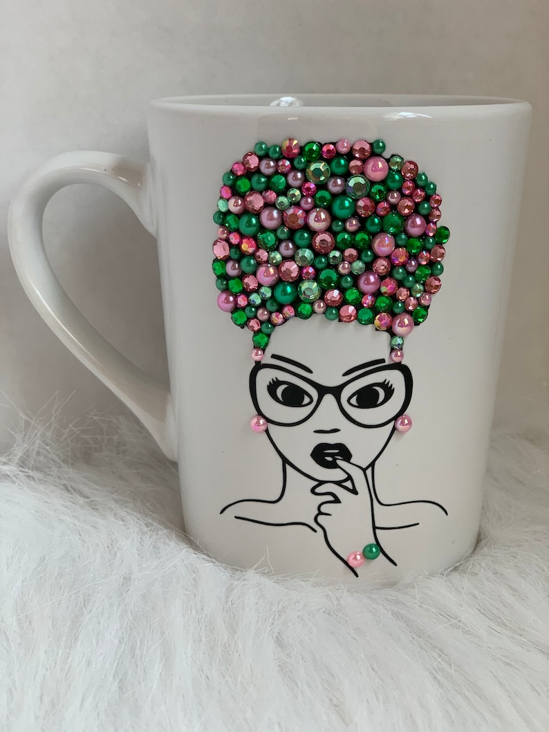 May include: White ceramic mug with a black line drawing of a woman with glasses and an afro. The afro is decorated with pink and green rhinestones.
